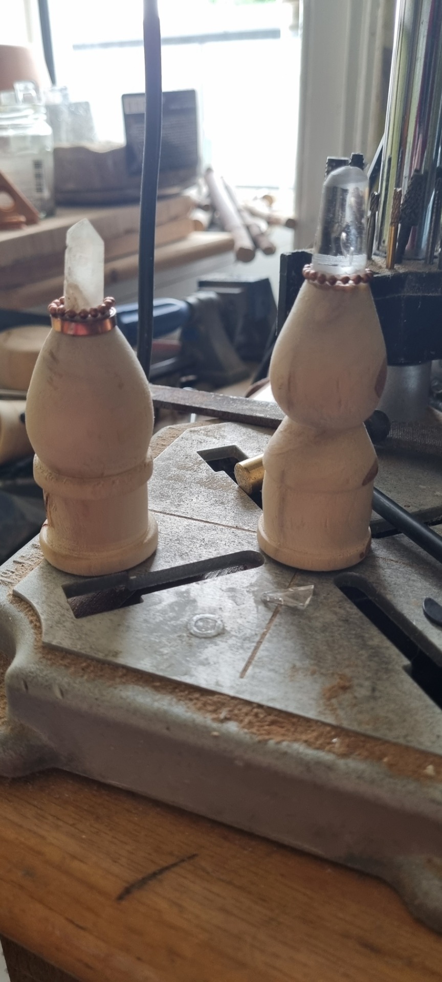 Two carved wooden pendulums with copper beaded bands on a metal workbench, workshop background blurred.